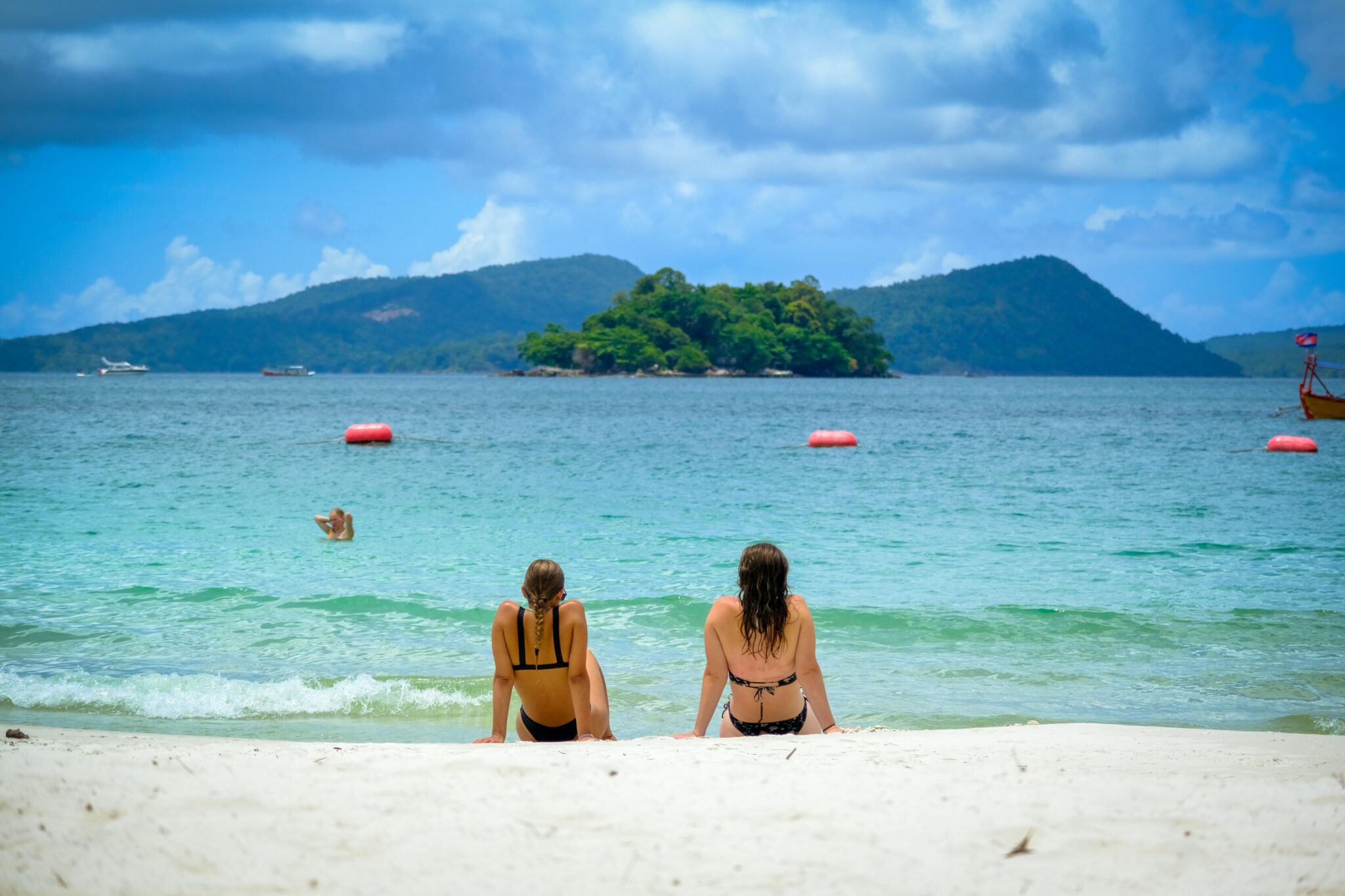 Two women enjoying the sunny beach in Sihanoukville, Cambodia, with clear blue water and a scenic island view.